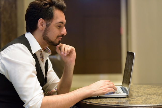 Vista di profilo del giovane uomo indiano bello pensare mentre si utilizza il computer portatile nella hall dell'hotel