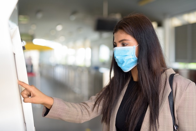 Profile view of young Indian businesswoman with mask buying ticket at skytrain station