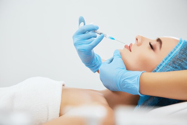 Profile of woman in blue cap lying on couch in cosmetological clinic with closed eyes. Doctor's hands in blue gloves doing lips augmentation with syringe with filler, closeup