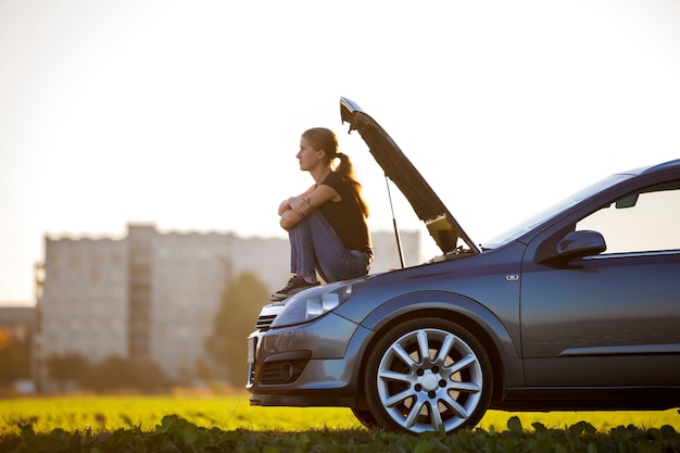 Profile of young slim attractive woman sitting on car with popped hood in green meadow waiting for help on clear sky copy space background