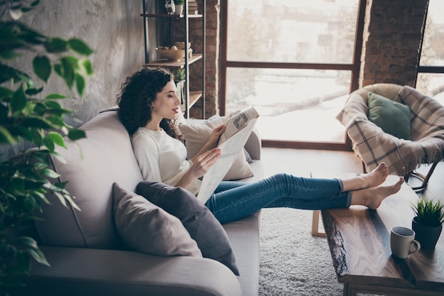 Photo profille side view portrait of     charming peaceful   girl sitting on sofa reading day finance news at modern industrial loft interior style living-room indoors