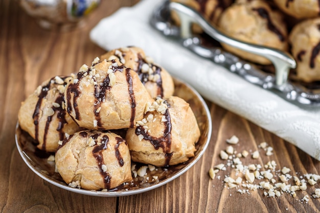 Profiteroles is a delicious culinary dishes of French cuisine with chocolate icing and nuts. Delicious profiteroles dessert on plate on dark wooden background.