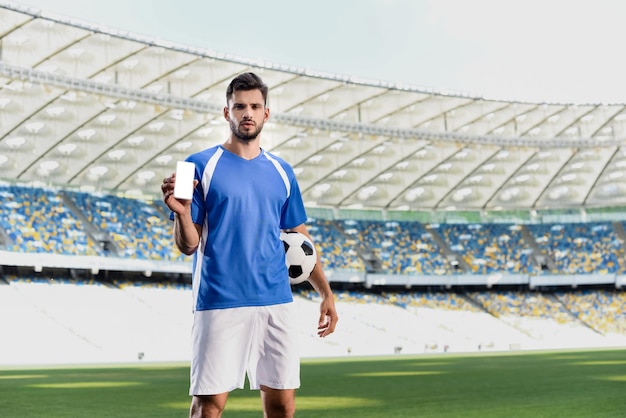 Profvoetballer in blauw en wit uniform met bal met smartphone met leeg scherm