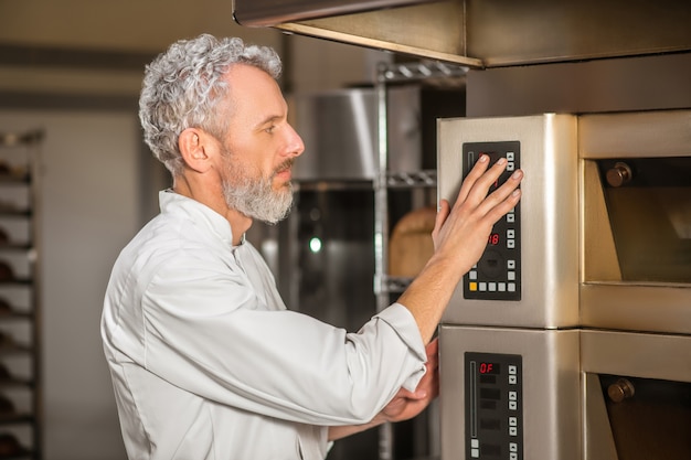 Program selection, oven. Profile of serious gray-haired bearded man in white jacket near control panel of stove touching with hand choosing program