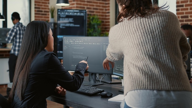 Programer pointing pencil at computer screen with software compiling code explaining source algorithm to coworker standing next to desk. Programmers discussing artificial intelligence with team.