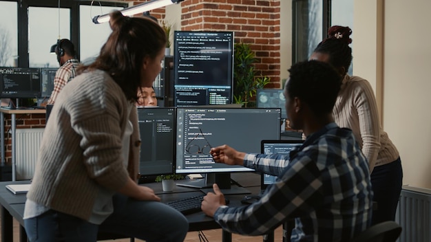 Programer sitting on desk discussing with mixed team of software developers about artificial intelligence innovation. Programmers doing teamwork looking at running machine learning algorithms.