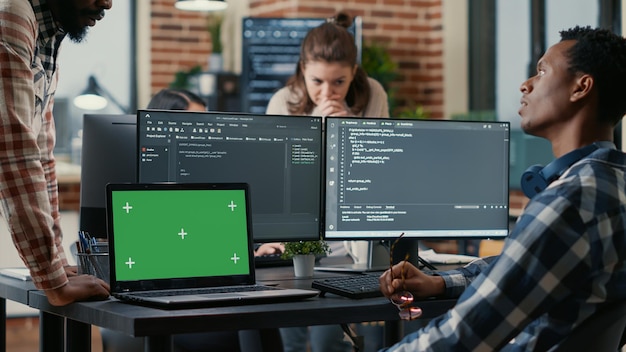 Programer taking glasses off talking with colleague at desk\
with laptop with green screen chroma key mockup and multiple\
computer screens compiling running code. coders doing online cloud\
computing.