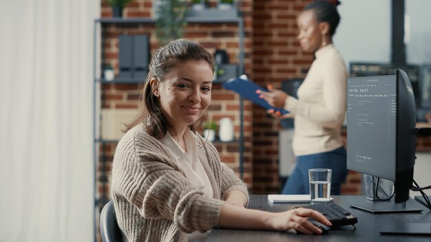 Programer typing source code on computer keyboard turning head and smiling at camera. Casual software engineer sitting at desk in it startup company writing ai algorithm feeling confident.