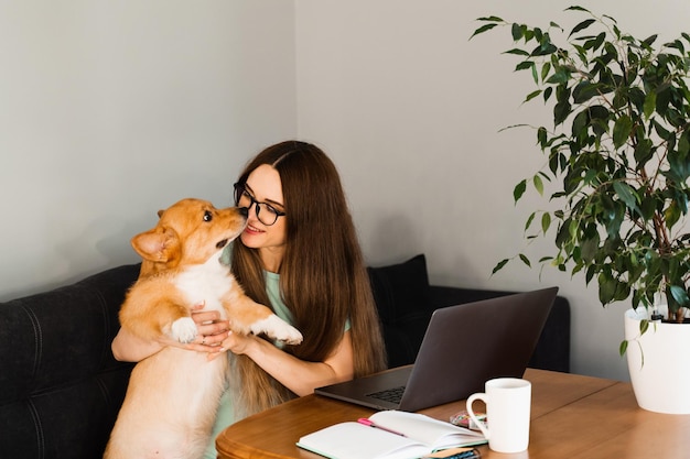 안경을 쓴 프로그래머 소녀는 노트북 작업을 하고 집에서 Welsh Corgi Pembroke 강아지와 즐거운 시간을 보내며 휴식을 취합니다. IT 전문가 여성은 사랑스러운 강아지를 안고 쓰다듬어줍니다