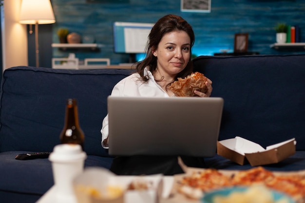 Programmer relaxing on couch holding laptop eating a tasty\
takeaway burger and fast food delivery menu in living room. coder\
after work watching online series while having a hamburger.