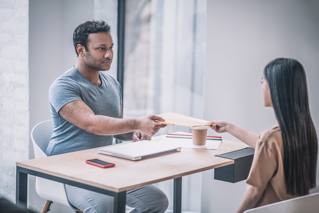 Progress report. Serious girl with long dark hair gives folder with documents to dark-skinned athletic guy sitting opposite each other at table in office