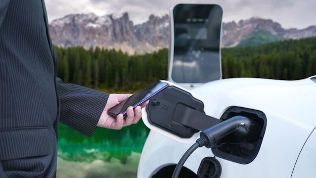 Progressive businessman checking EV car's battery status from smartphone at charging station with rural mountain background Combination of modern technology with natural scene Ecofriendly EV car