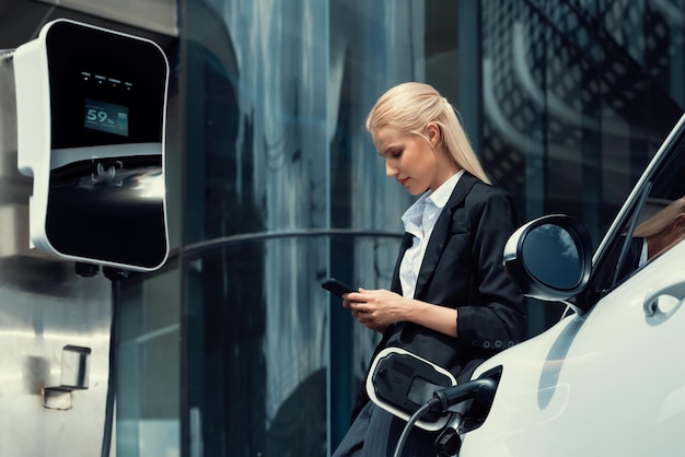 Прогрессивная деловая женщина с макетом смартфона с электромобилем на зарядной станции