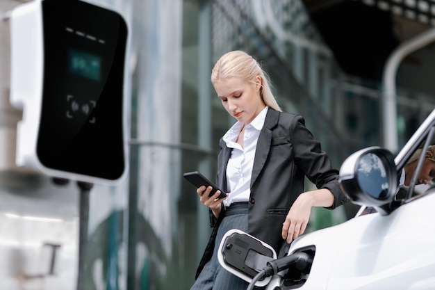 Progressive businesswoman with mockup smartphone with EV at charging station