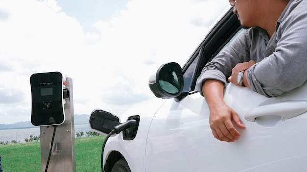 Progressive man connect power cable plug from charging station to his electric car EV car driven by green energy on green field for future clean environment concept