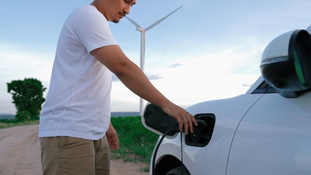 Progressive man with his electric car EV car recharging energy from charging station on green field with wind turbine as concept of future sustainable energy Electric vehicle with energy generator