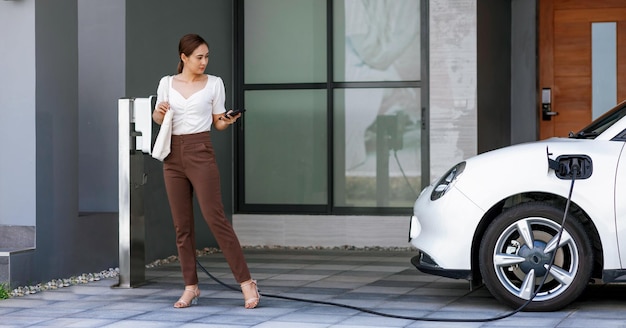 Progressive woman holding phone at EV home charging station in garage