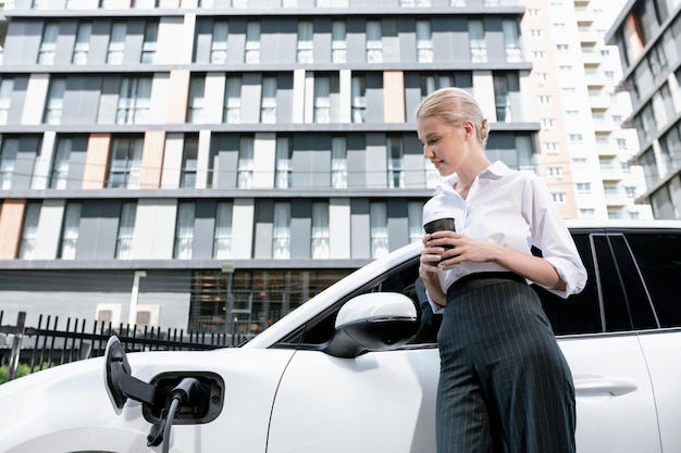 Progressive woman with coffee while charging EV car with residential buildings