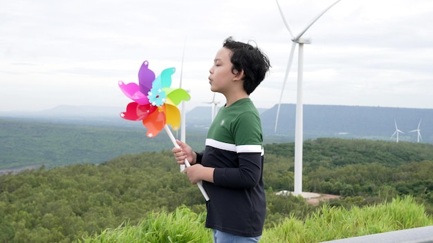 Photo progressive young asian boy playing with wind turbine toy in the wind turbine farm green field over the hill green energy from renewable electric wind generator