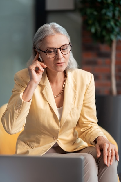 Project discussion. Beautiful grey-haired Caucasian woman with a wireless headset participating in a project discussion sitting in front of a laptop