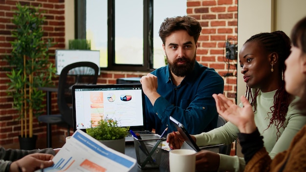 Project manager showing business charts on laptop to coworkers, talking about collaboration with data charts and research. Team of colleagues working on presentation report in office.