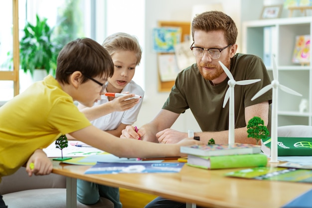 Projective poster. Concentrated bearded teacher spending time with his curious pupils in equipped bright classroom