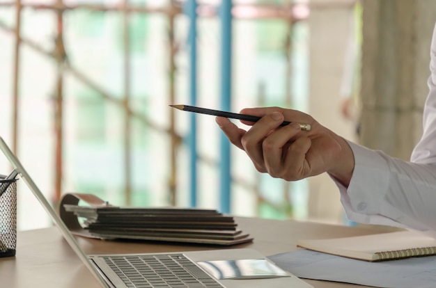 Projectmanagerhand die met pen op laptop computer en blauwdruk op bureau richten tijdens het bespreken van schaven voor nieuw project bij bouwwerf, contractant, techniek en bouwconcept