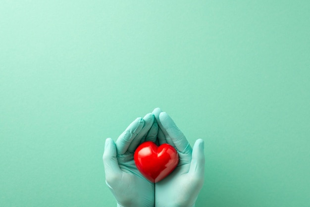 promote heart disease prevention with this topdown image of gloved hands holding a heart over a teal isolated background with ample copyspace available for text or advertisements