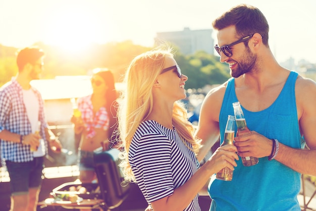 Proost op ons! Glimlachend jong koppel rammelende glazen met bier en kijken naar elkaar terwijl twee mensen barbecueën op de achtergrond