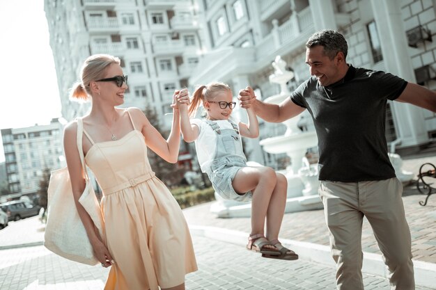 Prop and stay. Little smiling girl holding hands of her cheerful parents and hanging on them walking in the street.