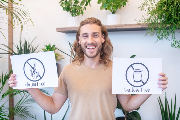 Proper nutrition. Joyful young bearded man in t-shirt holding showing gluten free lactose free posters standing indoors