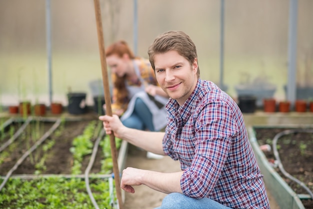 Prosperity. Friendly smiling confident young man with rake crouched near seedling bed and woman working in back of greenhouse