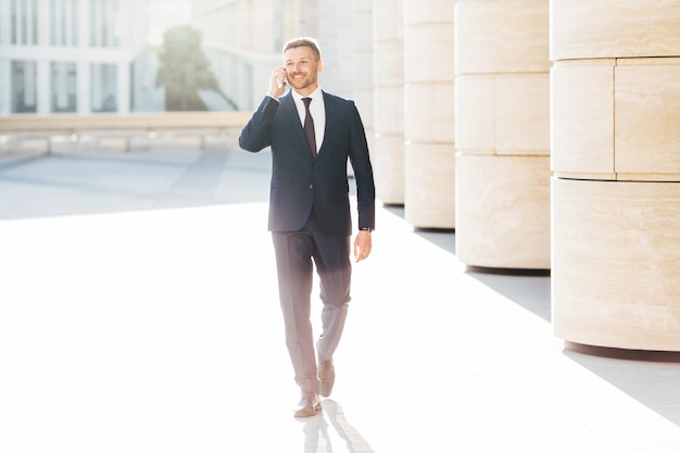 Prosperous male business owner in formal suit, has telephone conversation with business partner