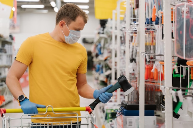 Protection from coronavirus Man chooses ax in building department of shopping mall poses with shopping cart wears disposable rubber gloves and mask prevents spread covid19 Pandemic outbreak