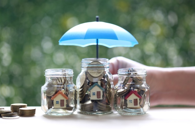 Protection,Model house and gold coin money in the glass bottle with hand holding the umbrella on natural green background, Finance insurance and Safe investment concept