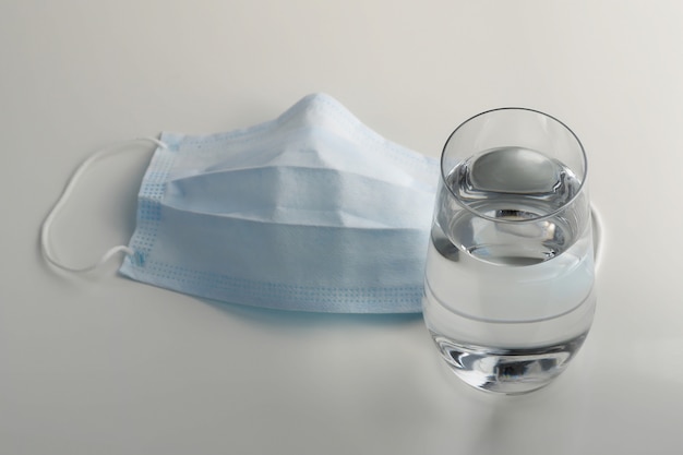 Maschera protettiva e una tazza di vetro con acqua limpida su un tavolo bianco. maschera medica per prevenire il coronavirus (covid-19).