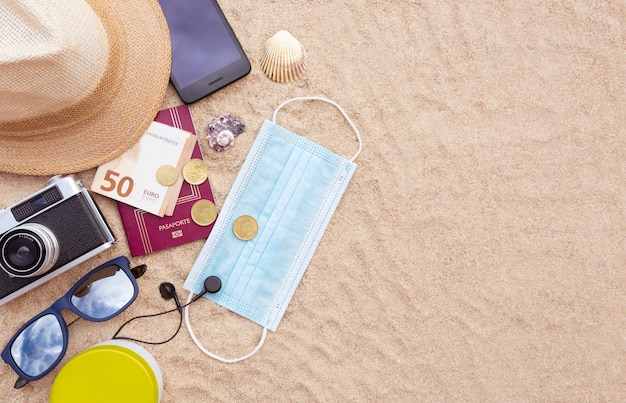 protective face mask, a passport and some cash, a smartphone, hat, camera and sunglasses on the beach sand. Flat lay