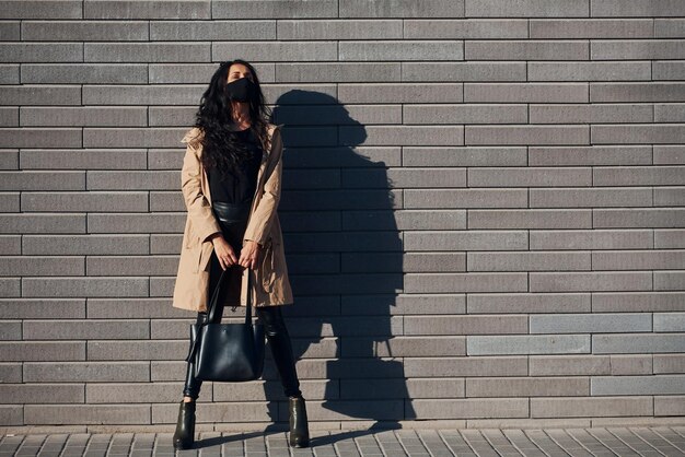 In protective mask Beautiful brunette with curly hair and in black clothes outdoors near wall