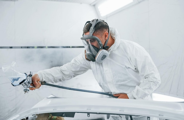 In maschera protettiva dipingere l'auto il riparatore di automobili caucasico in uniforme lavora in garage