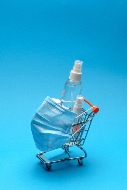 Protective masks and antibacterial disinfection sanitizer gel in a small shopping trolley cart on blue background with copy space.