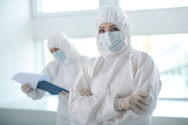 Protective measures. female medical worker in protective\
clothing and medical mask standing with folded arms, her colleague\
standing behind with clip folder