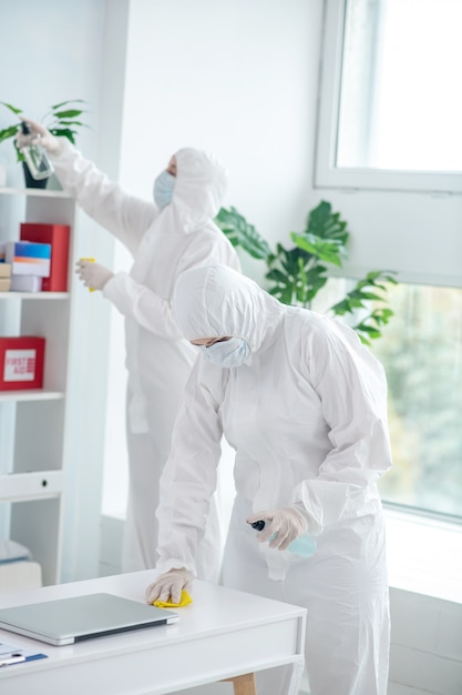 Protective measures. Medical worker in protective clothing and medical mask disinfecting table, another worker standing behind, cleaning closet