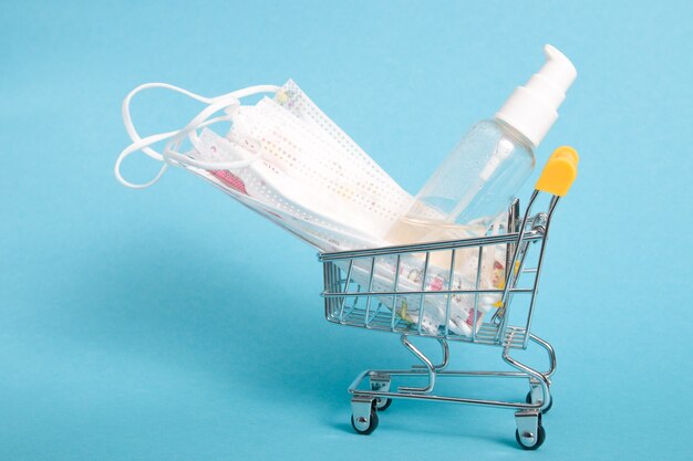 Protective medical face masks and sanitizer gel in a small shopping trolley, blue background, copy space