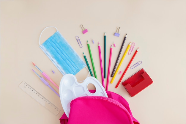 Foto una maschera medica protettiva e materiale scolastico e per ufficio sparsi da uno zaino rosa.