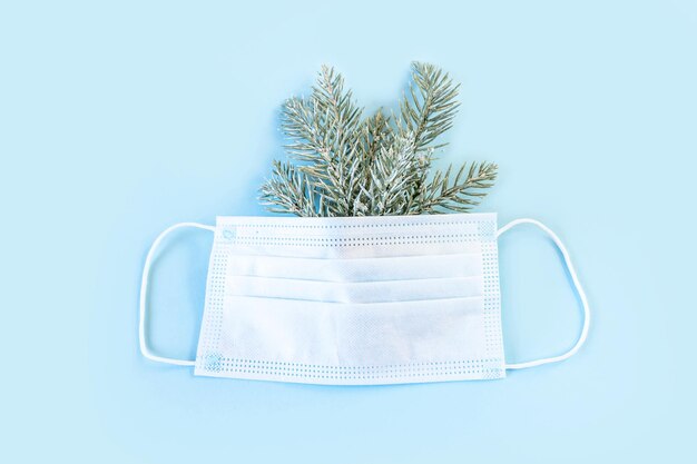 Protective medical mask with spruce branches on blue background. Christmas and New Year during pandemic of coronavirus infection. Flat lay, top view, copy space.