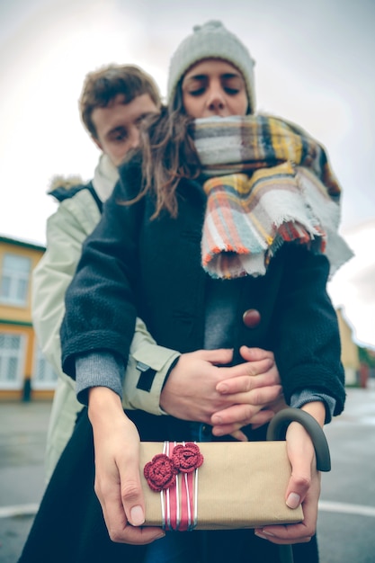 Protrait of young beautiful woman showing gift box in her hands and her boyfriend embracing from behind outdoors in a cold autumn day. Love and couple relationships concept.