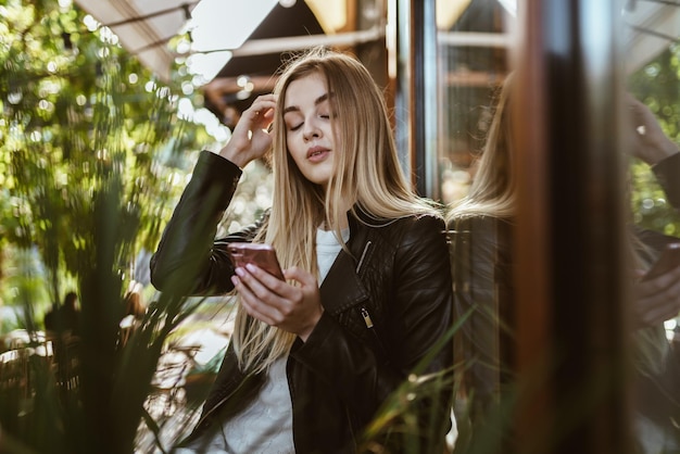 A proud beauty with long blonde hair is standing at the cafe and lowering her eyes straightens her hair with her hand in her other hand the phone