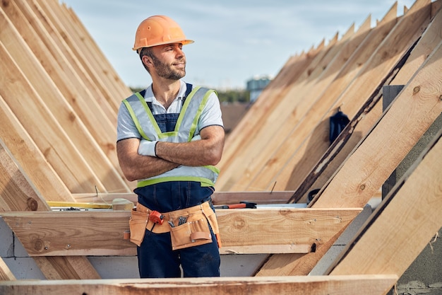 Costruttore orgoglioso con un elmetto che sembra soddisfatto del suo lavoro mentre è in piedi con le braccia incrociate in un cantiere