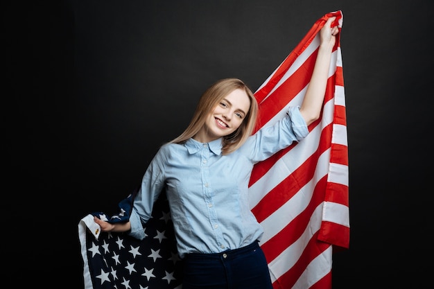 Proud cheerful happy girl having fun and smiling while holding national flag and standing isolated in black wall