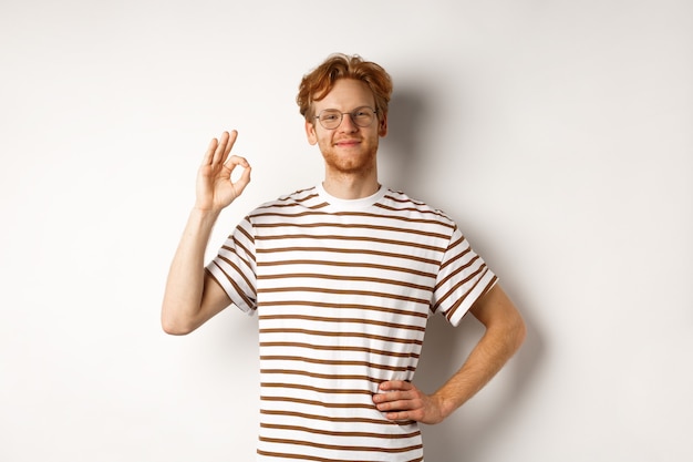 Proud and happy man with red hair and glasses smiling, showing OK sign, praising something excellent, saying yes or good, standing over white background.